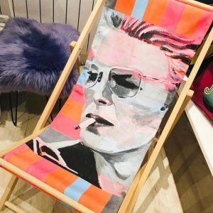 A neon bowie deck chair with a wooden frame, and a canvas sling seat with a David Bowie design printed on it.