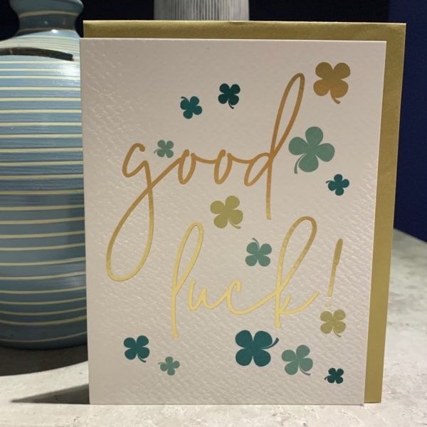 A card with the words Good Luck printed on it and lots of different coloured green four leaf clovers printed around the wording,