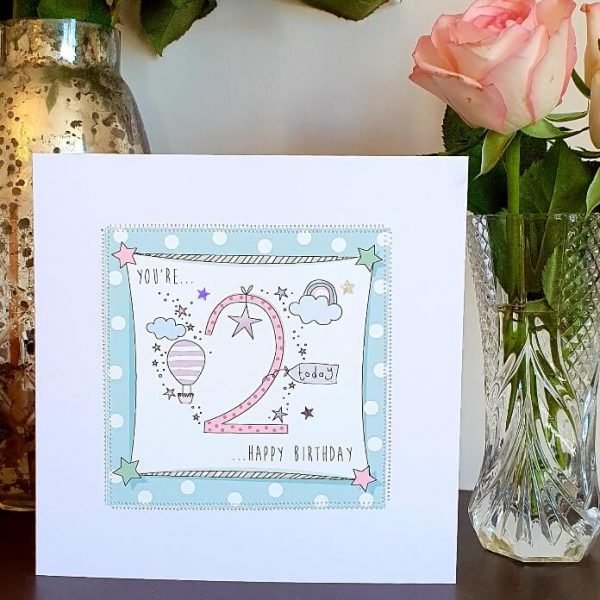 A 2nd birthday card in pinks and blues with a big pink spotty 2 and rainbows, stars and balloons. Hand finished with stitching and silver stars