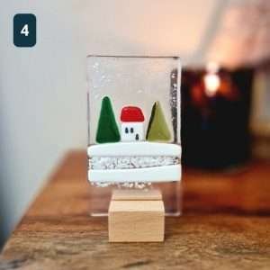 A fused glass Christmas ornament with trees, snow and a little house. On a wooden stand