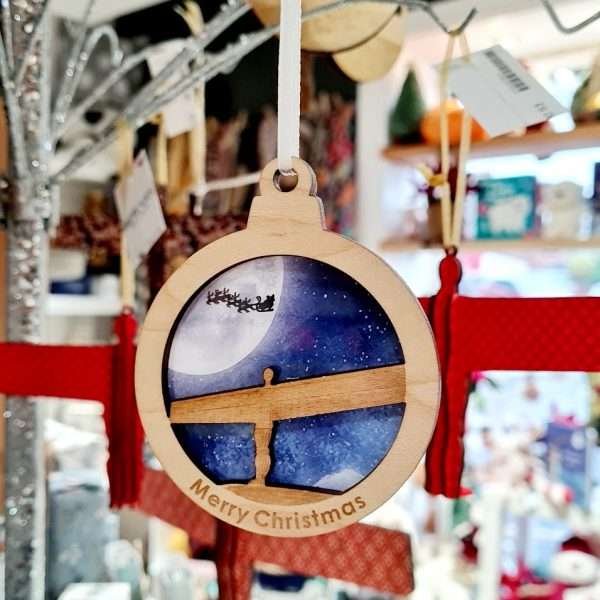 A laser cut wooden christmas bauble with the angel of the north set against a big moon sky with santa and his reindeer flying over