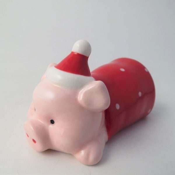 A ceramic pig lying down in a red and white spotty blanket wearing a red santa hat